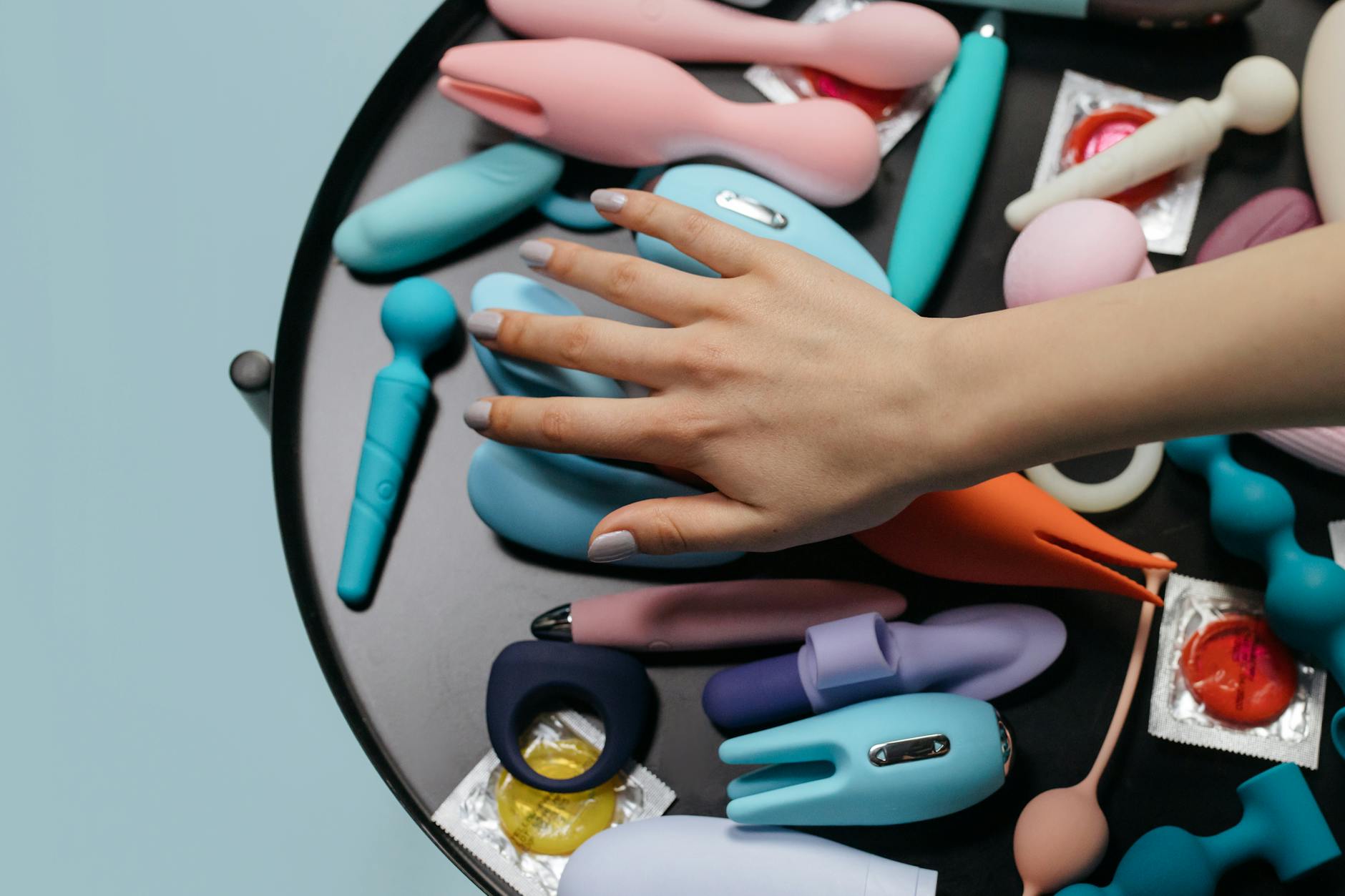Hand reaching over a variety of colorful sex toys arranged on a table.